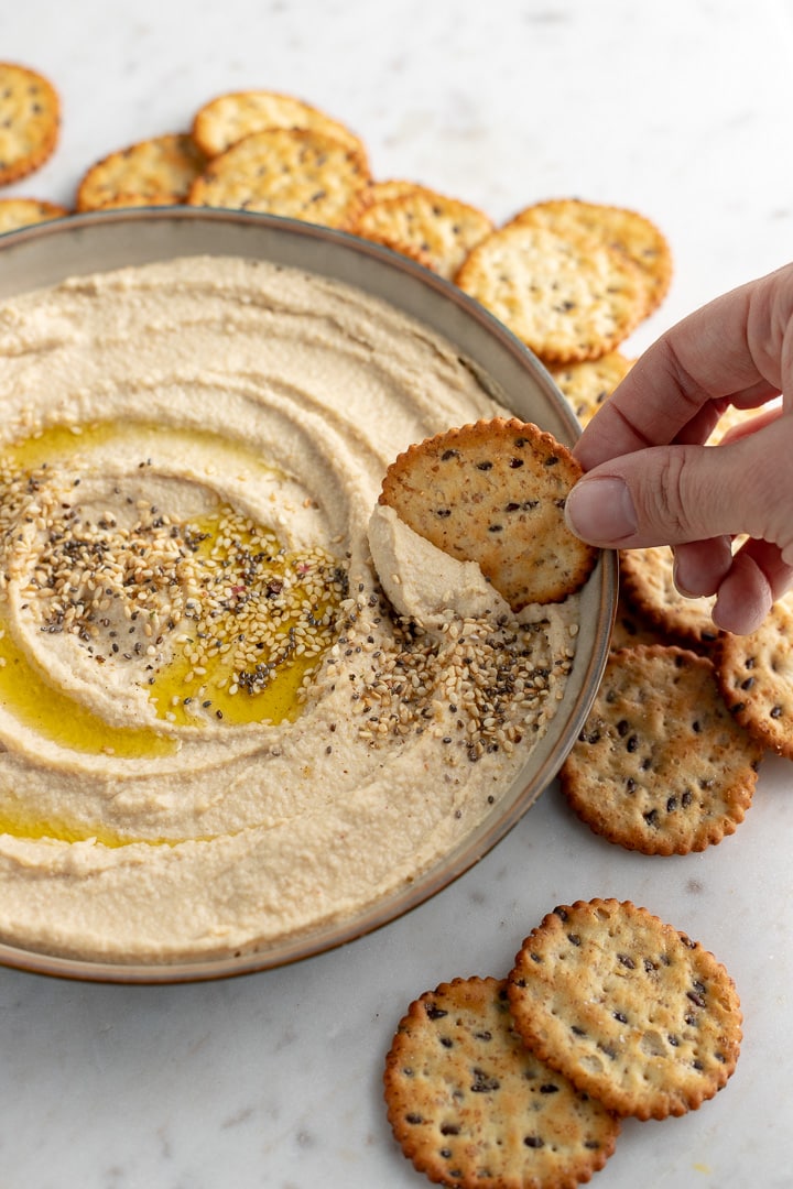 A hand dipping a cracker into a bowl of homemade hummus.