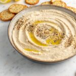 Serving bowl filled with smooth, creamy homemade hummus, garnished with olive oil and sesame seeds, surrounded by crackers.