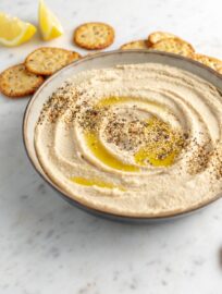 Serving bowl filled with smooth, creamy homemade hummus, garnished with olive oil and sesame seeds, surrounded by crackers.
