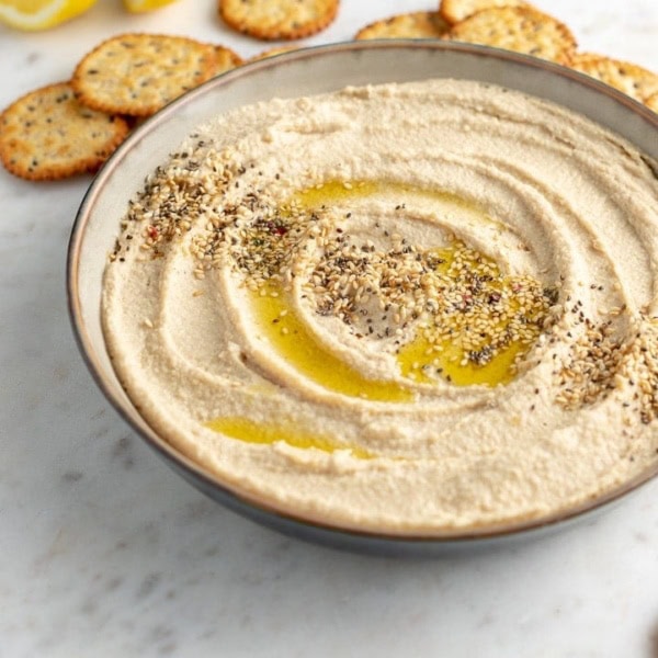Serving bowl filled with smooth, creamy homemade hummus, garnished with olive oil and sesame seeds, surrounded by crackers.