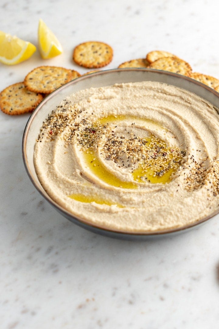 Close-up of a bowl of creamy homemade hummus, garnished with olive oil and sesame seeds, surrounded by crackers.