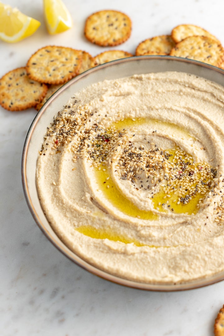 Serving bowl filled with smooth, creamy homemade hummus, garnished with olive oil and sesame seeds, surrounded by crackers.