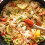 Close up of a serving spoon lifting a helping of lemon shrimp orzo out of a cast iron skillet.