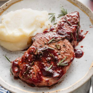 Plate with a serving of mashed potatoes and a balsamic plum pork chop garnished with fresh rosemary.