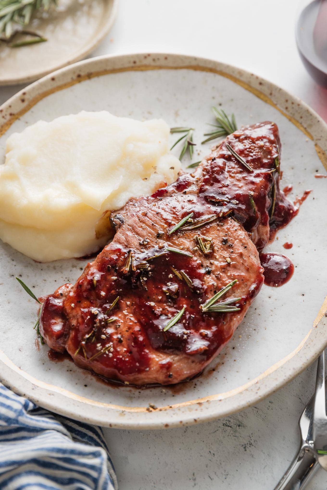 Plate with a serving of mashed potatoes and a balsamic plum pork chop garnished with fresh rosemary.