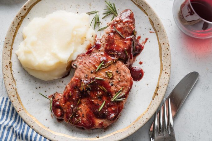 Landscape oriented overhead photo of a plate with a pork chop in balsamic plum sauce served with mashed potatoes and a glass of red wine nearby.