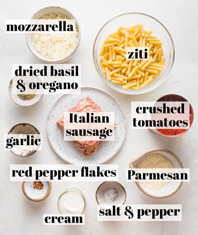 Labeled overhead image of ground Italian sausage, ziti, crushed tomatoes, dried basil, dried oregano, garlic cloves, mozzarella, Parmesan, cream, salt, and pepper, all measured into prep bowls and ready to cook.