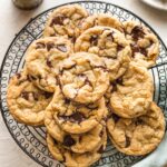 Bakery style chocolate chip cookies stacked on a black metal cooling rack with glasses of milk around it.