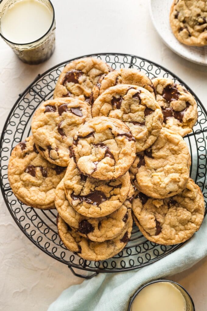 Bakery style chocolate chip cookies stacked on a black metal cooling rack with glasses of milk around it.