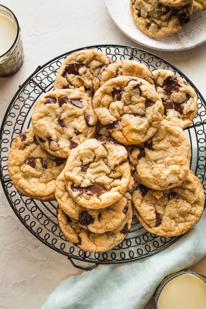 Bakery style chocolate chip cookies stacked on a black metal cooling rack with glasses of milk around it.