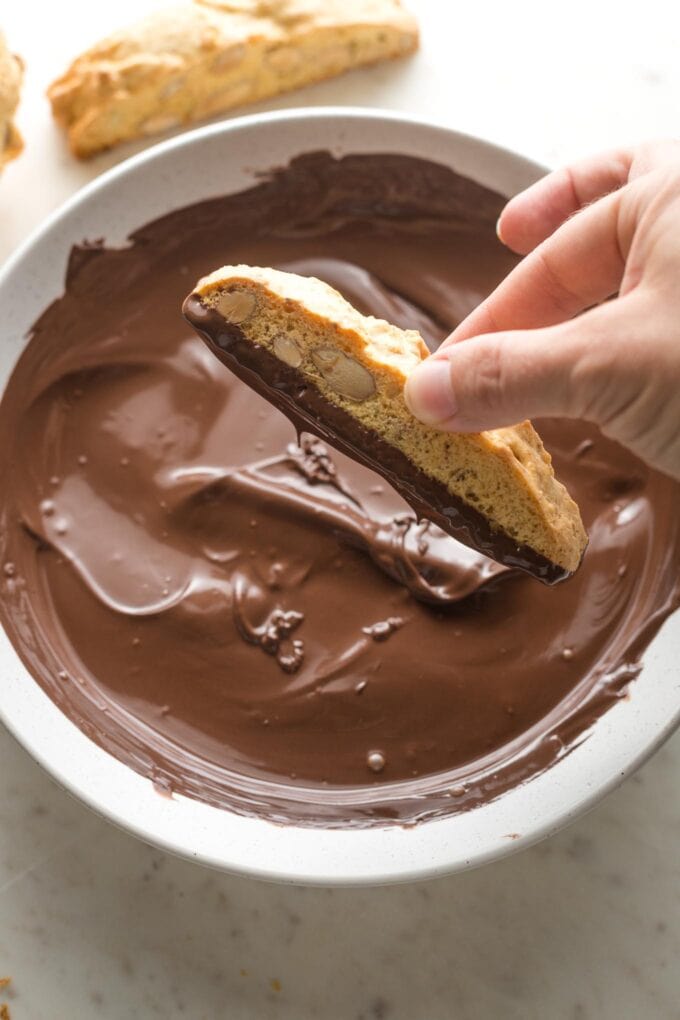 Hand dipping a baked biscotti cookie into melted chocolate.