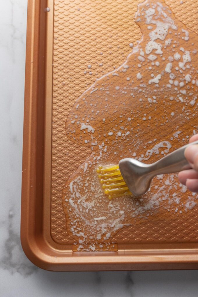 Pastry brush spreading melted butter over a rimmed baking sheet.