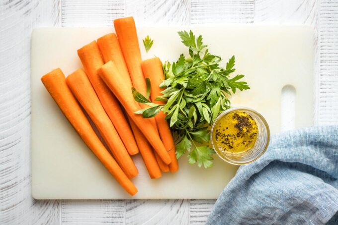 Peeled carrots, flat-leaf parsley, and a jar of salad dressing on a white cutting board.