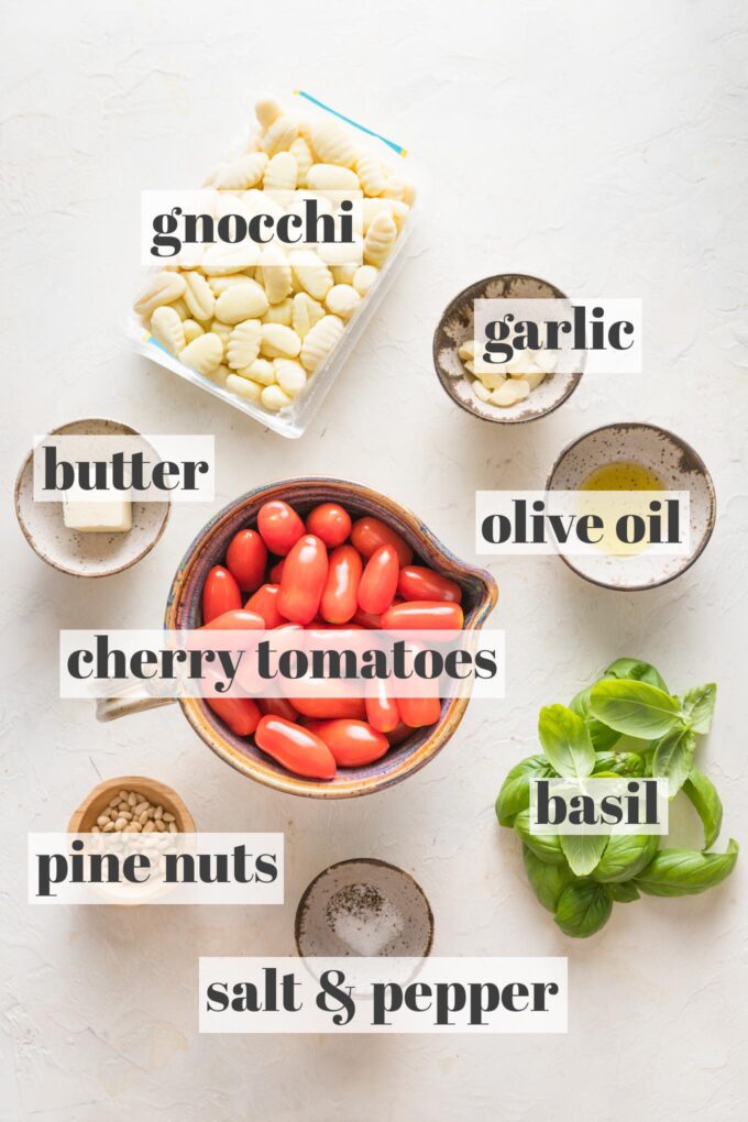 Labeled overhead image of cherry tomatoes, potato gnocchi, garlic, olive oil, butter, fresh basil, pine nuts, salt, and pepper in prep bowls.