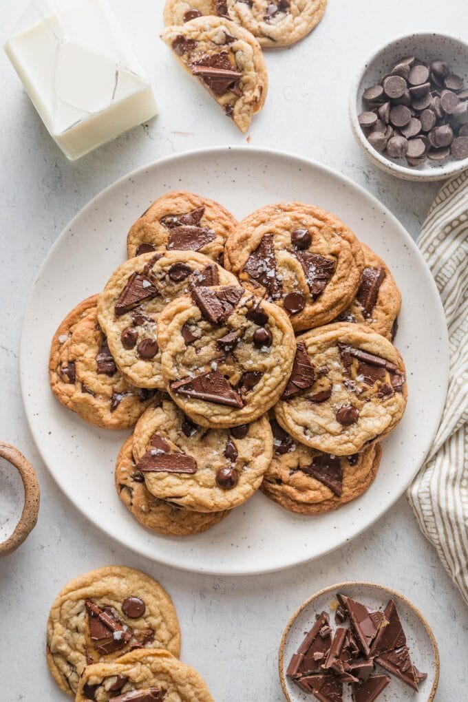 Countertop set with a large platter of salted chocolate chip cookies, with extra chocolate caramel squares, chocolate chips, and a jar of milk scattered to the edges.