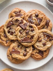 Overhead view of a speckled white plate piled high with salted caramel chocolate chip cookies, with extra chocolate chips and a small glass bottle of milk resting off to the side.