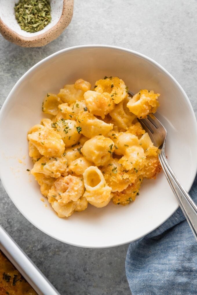 Close up of a fork in a bowl of creamy baked mac and cheese served in a cozy white bowl with breadcrumb topping and parsley.