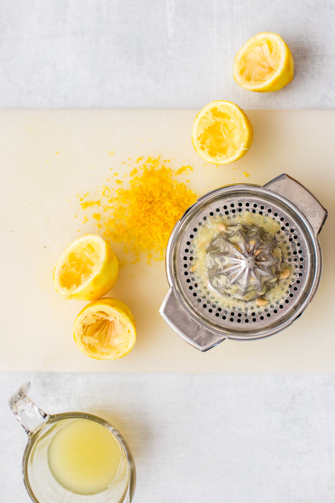 Scattered, juice lemons, a hand juicer, and a small bowl of lemon zest, on a light white cutting board.