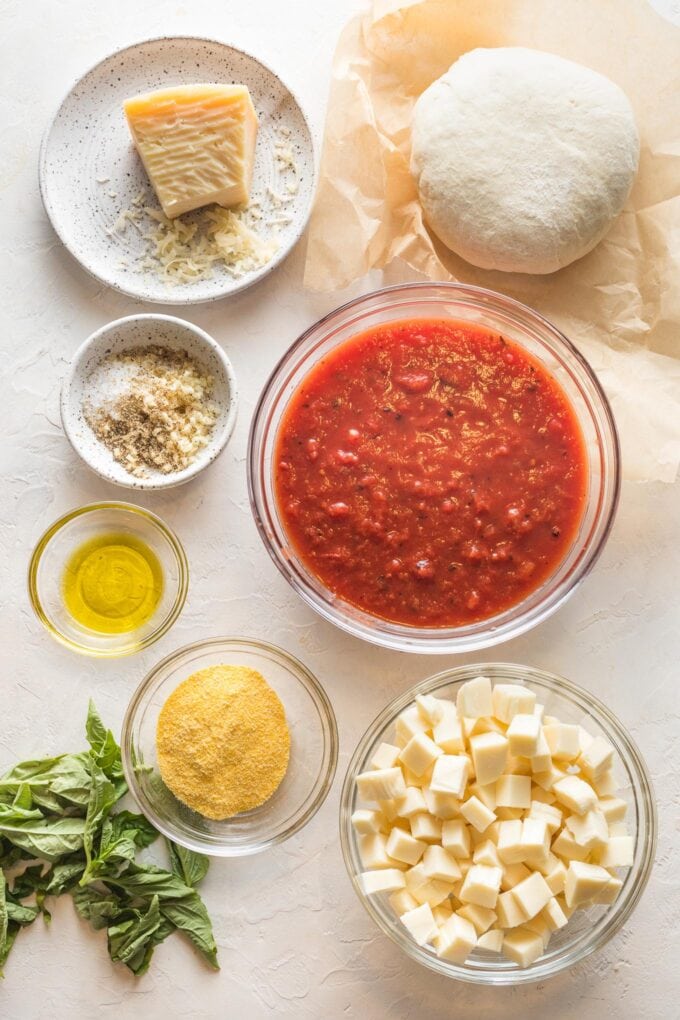 A ball of pizza dough and prep bowls holding crushed tomatoes, seasonings, olive oil, cornmeal, Parmesan, and mozzarella cheese.