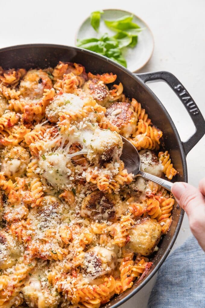 Serving spoon lifting up a generous helping from a large pan of baked chicken Parmesan pasta.