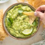 Hand dipping a crisp tortilla chip into a bowl of homemade guacamole.