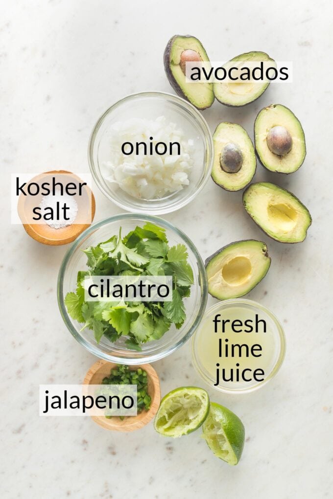 Sliced avocados and prep bowls holding chopped onion, kosher salt, cilantro, lime juice, and diced jalapeno pepper.