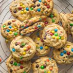 Cooling rack full of chocolate chip M&M cookies with a nearby glass of milk.