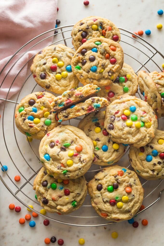 Cooling rack full of chocolate chip M&M cookies with one broken open to show the soft, chewy inside.