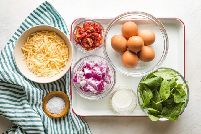 Prep bowls holding eggs, spinach, Parmesan and Gouda cheese, chopped red onion, sun-dried tomatoes, cream, salt and pepper.