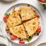 A black bean quesadilla with roasted red peppers cut into four wedges and served with avocado and salsa.