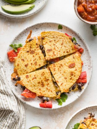 A black bean quesadilla with roasted red peppers cut into four wedges and served with avocado and salsa.