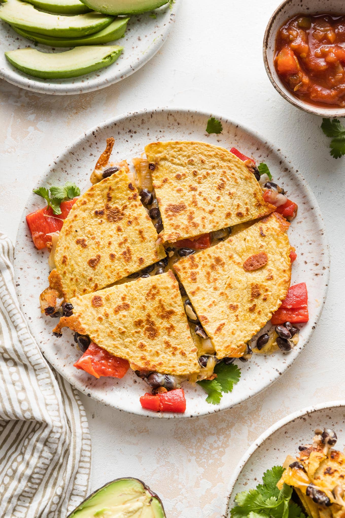 A black bean quesadilla with roasted red peppers cut into four wedges and served with avocado and salsa.