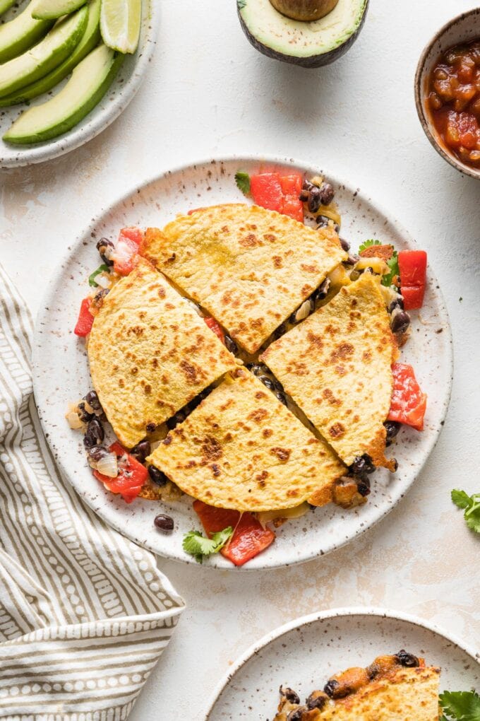 A black bean quesadilla with roasted red peppers cut into four wedges and served with avocado and salsa.