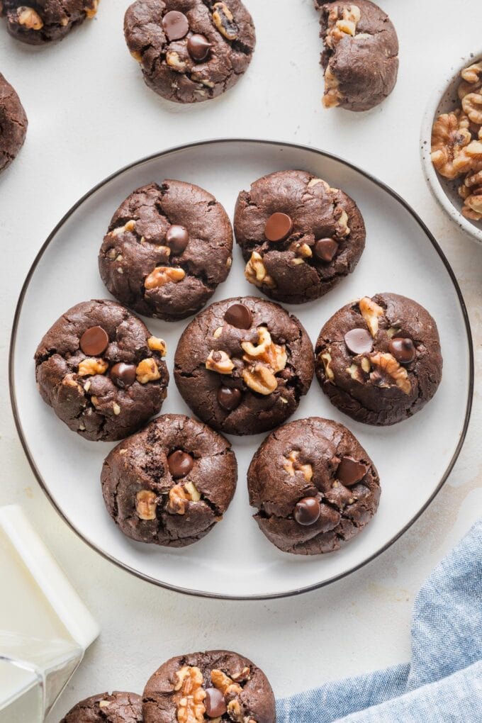 Small black-rimmed white plate holding seven double chocolate espresso walnut cookies, with a small carafe of milk and extra walnut halves in the background.