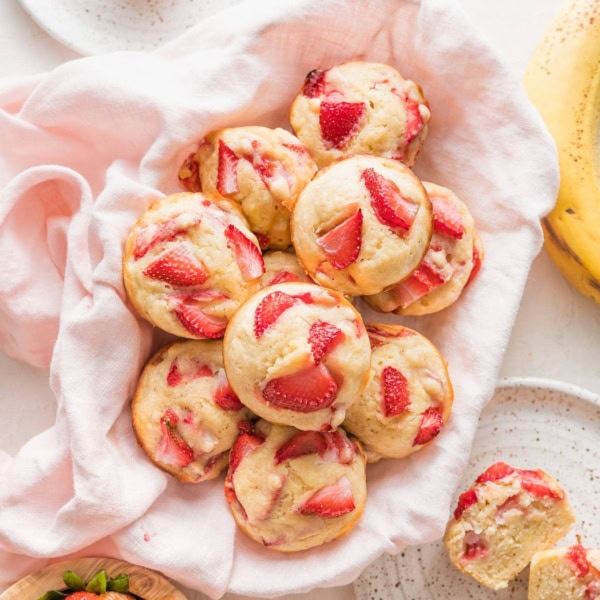 Basket filled with strawberry banana muffins, surrounded by small plates and extra fresh strawberries.