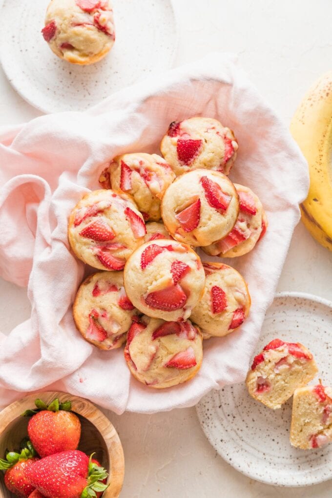 Basket filled with strawberry banana muffins, surrounded by small plates and extra fresh strawberries.