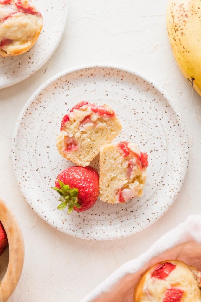 A small white plate with a sliced open strawberry banana muffin, cut side up, and one fresh strawberry.