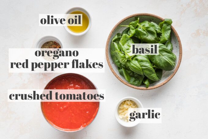 Labeled photo of crushed tomatoes, minced garlic, fresh basil leaves, olive oil, dried oregano, and red pepper flakes.