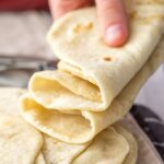 A hand holding a stack of soft, pliable homemade flour tortillas.