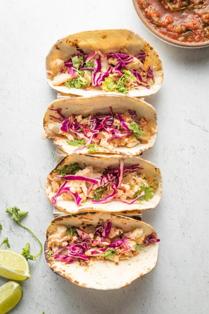 Four jalapeno lime fish tacos with shredded cabbage, lined up in a row.