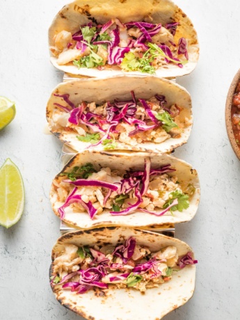 Four jalapeno lime fish tacos with shredded cabbage, lined up in a row.