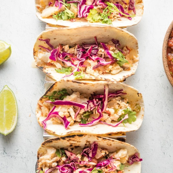 Four jalapeno lime fish tacos with shredded cabbage, lined up in a row.