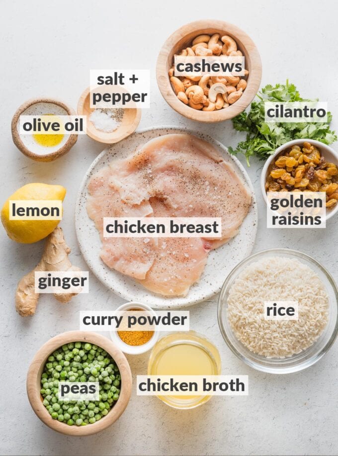 Labeled overhead photo of chicken breasts, rice, cashews, golden raisins, fresh cilantro, fresh ginger, a lemon, curry powder, peas, salt, and pepper, all measured into bowls and ready to cook.