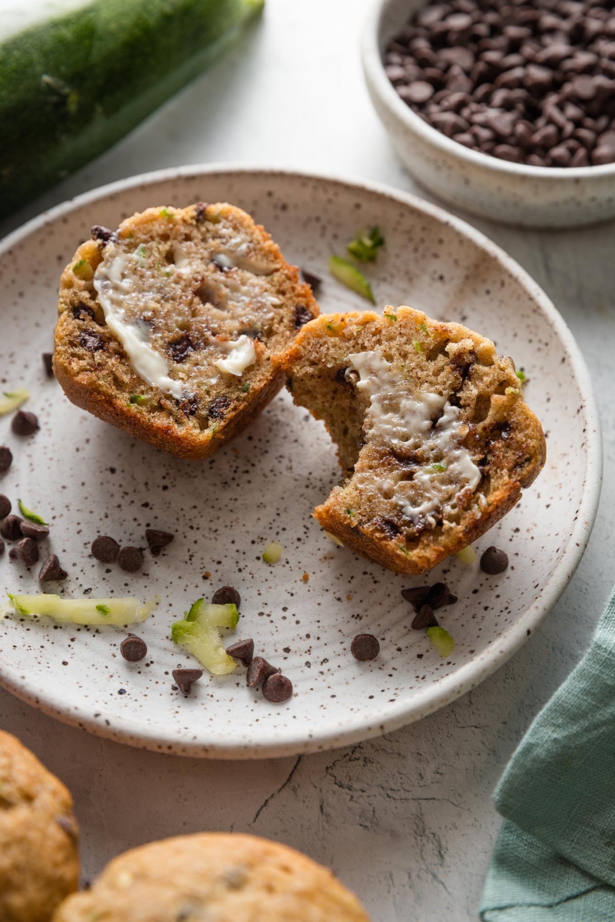 Small plate with a homemade zucchini chocolate chip muffin, split open to show the melty mini chocolate chips inside and slathered with a pat of partly melted butter.
