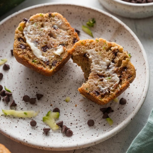 Small plate with a homemade zucchini chocolate chip muffin, split open to show the melty mini chocolate chips inside and slathered with a pat of partly melted butter.