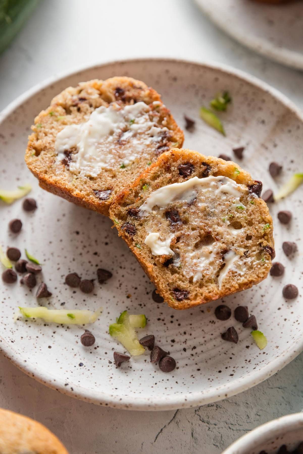 Small plate resting on a counter with a zucchini chocolate chip muffin on top, split open and topped with warm butter.