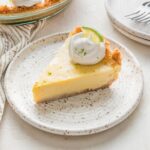 45 degree angle view of a slice of key lime pie on a white ceramic dessert plate, garnished with a dollop of whipped cream and sprinkle of lime zest.