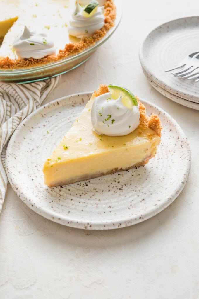 45 degree angle view of a slice of key lime pie on a white ceramic dessert plate, garnished with a dollop of whipped cream and sprinkle of lime zest.