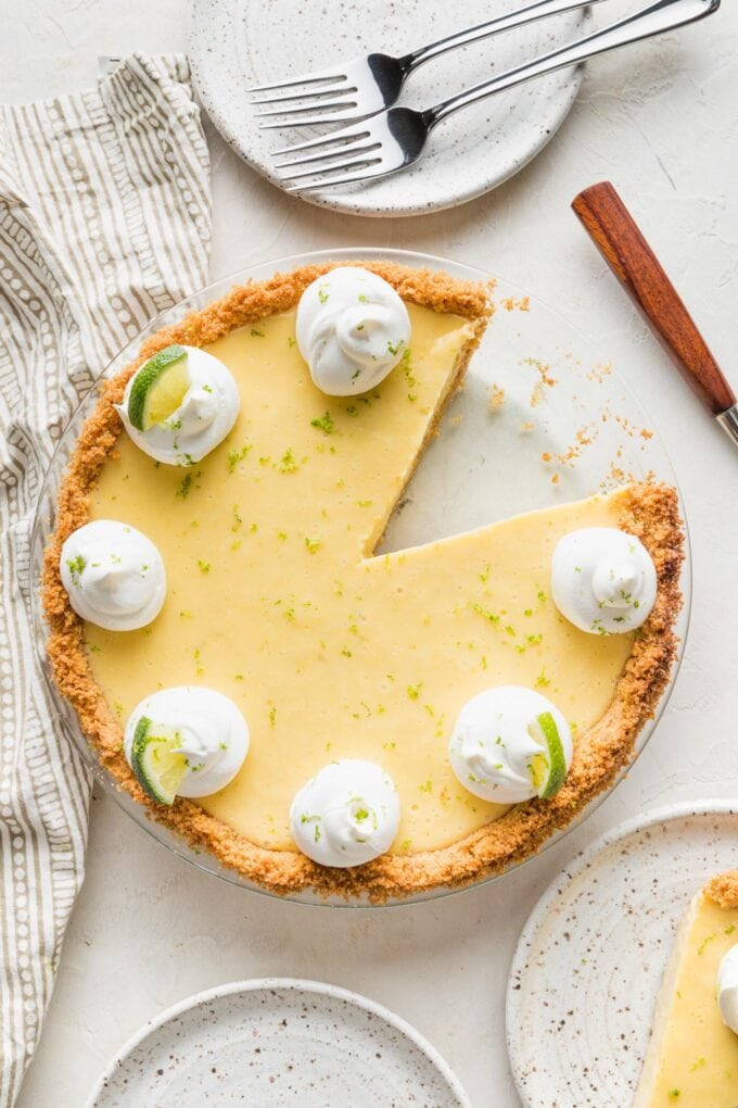 Overhead image of a key lime pie garnished with whipped cream, lime slices, and lime zest, with one slice cut out and served.