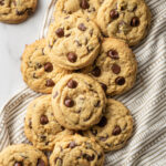 Pile of soft chocolate chip cookies on top of a napkin, with a glass of milk to the side.